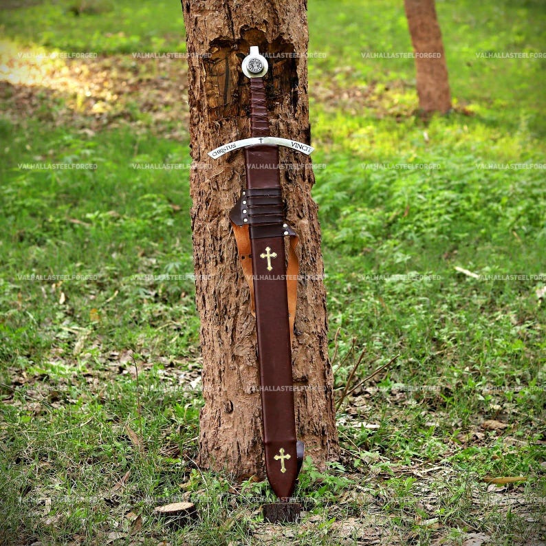 May include: A sword with a brown leather-wrapped hilt and a matching brown leather scabbard. The metal crossguard is engraved with the text CHRISTUS VINCIT. The scabbard features two gold-colored cross emblems, one near the top and one at the base.