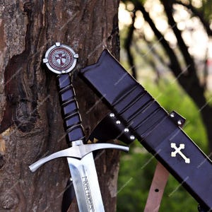May include: A steel sword with a black leather-wrapped grip and a circular pommel featuring a red cross emblem. The blade is engraved with the text NON NOBIS. Beside it is a black leather scabbard adorned with a gold-colored cross inlay.