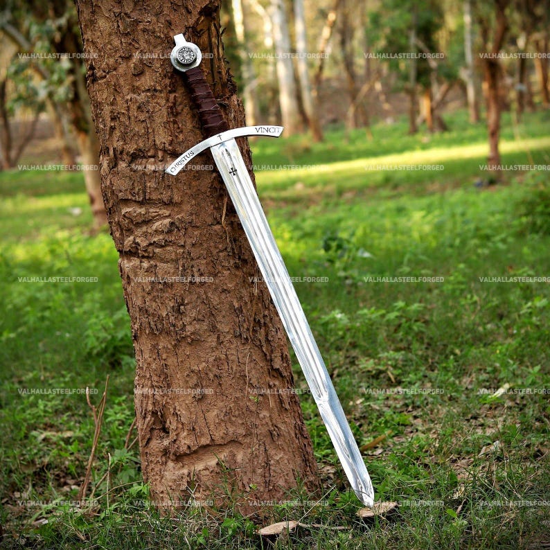 May include: A silver-toned steel sword with a dark brown leather-wrapped grip and a circular pommel. The crossguard features the engraved text "CHRISTUS" on one side and "VINCIT" on the other, with a small cross symbol centered on the blade.
