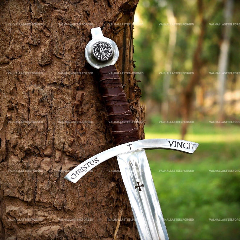 May include: A silver-toned metal sword featuring a brown leather-wrapped grip and a circular pommel with a circular emblem. The crossguard is engraved with the text CHRISTUS VINCIT and a cross symbol, with a small cross etched on the blade.