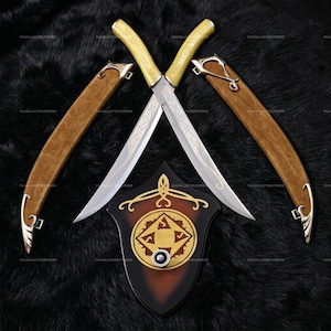 May include: Two crossed swords with silver blades featuring intricate etched scrollwork and pale yellow handles. Each sword has a matching tan leather scabbard with silver metal accents. Below the swords is a shield-shaped wall plaque.