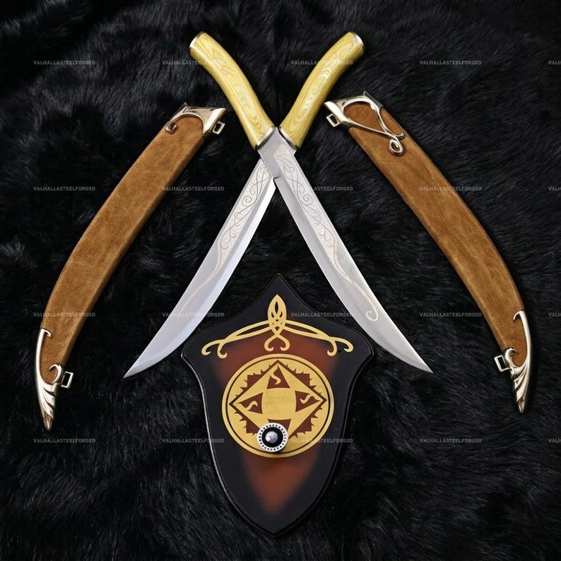 May include: Two crossed swords with silver blades featuring intricate etched scrollwork and pale yellow handles. Each sword has a matching tan leather scabbard with silver metal accents. Below the swords is a shield-shaped wall plaque.