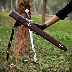 May include: A dark brown leather scabbard featuring two white cross emblems and a matching leather shoulder strap with small brass cross accents. The scabbard is designed to hold a sword, with a visible metal blade and hilt resting against a tree.