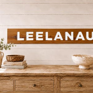 Könnte beinhalten: Ein rechteckiges Wand-Schild aus Holz mit dem Wort LEELANAU in fetten, weißen Großbuchstaben. Das Schild besteht aus einer einzelnen horizontalen Holzplanke in mittlerem Braunton mit sichtbarer Maserung und einem schlichten Design.