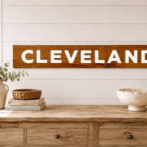 Könnte beinhalten: Ein rechteckiges Wand-Schild aus Holz mit dem Wort CLEVELAND in fetten, weißen Großbuchstaben, zentriert auf einer dunkelbraunen, horizontalen Holzplanke mit sichtbarer Maserung, montiert an einer weißen Wand mit horizontalen Paneelen.