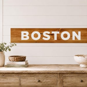 Könnte beinhalten: Ein rechteckiges Holzschild mit einer natürlichen braunen Holzmaserung, das das Wort BOSTON in großen, fetten, weißen Großbuchstaben zeigt, die horizontal in der Mitte der Vorderseite angebracht sind und an einer weißen Wand mit Paneelen hängen.