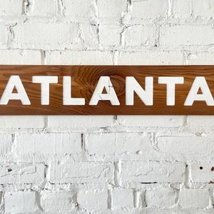 K&ouml;nnte beinhalten: Ein rechteckiges Holzschild mit einer nat&uuml;rlichen braunen Holzmaserung, auf dem das Wort ATLANTA in gro&szlig;en, fetten, wei&szlig;en Gro&szlig;buchstaben mittig angebracht ist. Das Schild hat eine glatte Oberfl&auml;che mit einem sichtbaren Astloch in der Maserung.