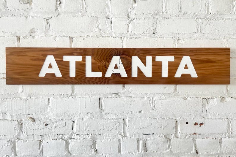 K&ouml;nnte beinhalten: Ein rechteckiges Holzschild mit einer nat&uuml;rlichen braunen Holzmaserung, auf dem das Wort ATLANTA in gro&szlig;en, fetten, wei&szlig;en Gro&szlig;buchstaben mittig angebracht ist. Das Schild hat eine glatte Oberfl&auml;che mit einem sichtbaren Astloch in der Maserung.
