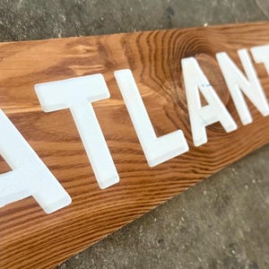 K&ouml;nnte beinhalten: Ein rechteckiges Holzschild mit einer nat&uuml;rlichen braunen Holzmaserung und dem Wort ATLANTA in fetten, erhabenen, wei&szlig;en Gro&szlig;buchstaben. Die Schrift ist auf dem horizontalen Brett zentriert und erzeugt einen kontrastreichen visuellen Effekt.
