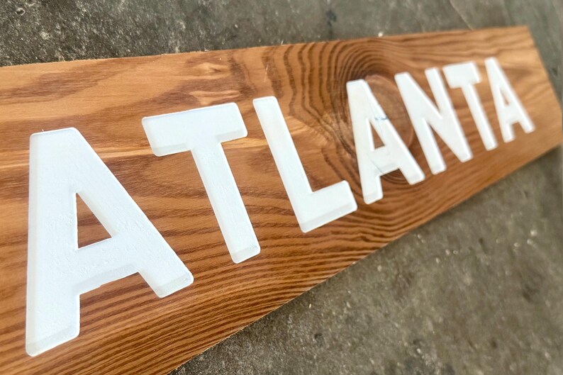 K&ouml;nnte beinhalten: Ein rechteckiges Holzschild mit einer nat&uuml;rlichen braunen Holzmaserung und dem Wort ATLANTA in fetten, erhabenen, wei&szlig;en Gro&szlig;buchstaben. Die Schrift ist auf dem horizontalen Brett zentriert und erzeugt einen kontrastreichen visuellen Effekt.