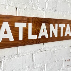 K&ouml;nnte beinhalten: Ein rechteckiges Holzschild mit einer nat&uuml;rlichen braunen Holzmaserung, auf dem das Wort ATLANTA in fetten, wei&szlig;en, erhabenen Gro&szlig;buchstaben zu sehen ist. Das Schild ist horizontal ausgerichtet und weist eine glatte Holzoberfl&auml;che auf.