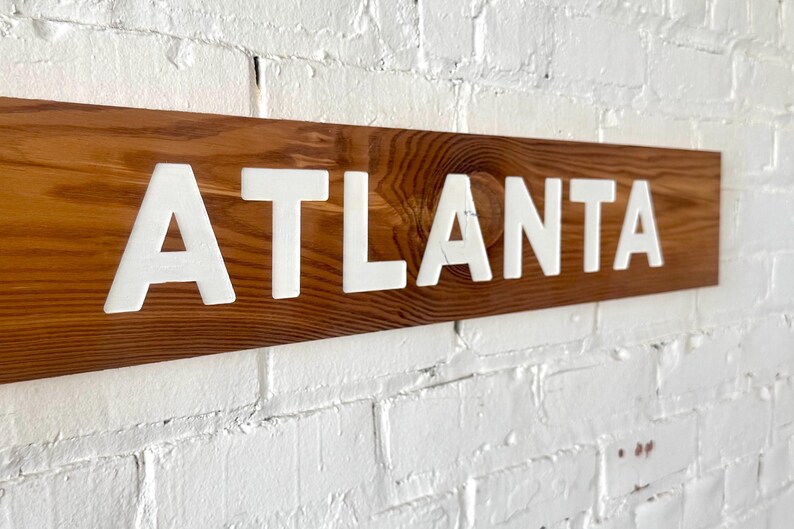K&ouml;nnte beinhalten: Ein rechteckiges Holzschild mit einer nat&uuml;rlichen braunen Holzmaserung, auf dem das Wort ATLANTA in fetten, wei&szlig;en, erhabenen Gro&szlig;buchstaben zu sehen ist. Das Schild ist horizontal ausgerichtet und weist eine glatte Holzoberfl&auml;che auf.