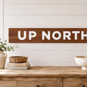 Könnte beinhalten: Ein rechteckiges Wand-Schild aus Holz mit einer dunkelbraunen Holzmaserung und der Aufschrift UP NORTH in fetten, erhabenen weißen Großbuchstaben. Das Schild ist horizontal ausgerichtet und besitzt eine glatte, flache Oberfläche.