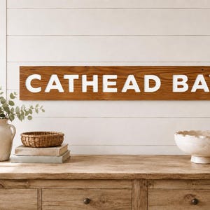 Könnte beinhalten: Ein rechteckiges Wand-Schild aus Holz mit dem Text CATHEAD BAY in fetten, weißen Großbuchstaben. Das Schild hat eine natürliche braune Holzmaserung und ist horizontal an einer weißen Wand mit Holzpaneelen befestigt.