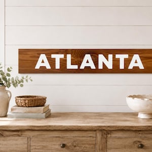 K&ouml;nnte beinhalten: Ein rechteckiges Holzschild mit dunkelbraun gebeizter Oberfl&auml;che und sichtbarer Holzmaserung. Das Wort ATLANTA ist in gro&szlig;en, fetten, wei&szlig;en Gro&szlig;buchstaben in der Mitte des horizontalen Brettes aufgedruckt und bildet einen klaren Kontrast.