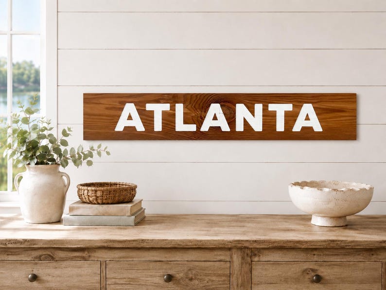 K&ouml;nnte beinhalten: Ein rechteckiges Holzschild mit dunkelbraun gebeizter Oberfl&auml;che und sichtbarer Holzmaserung. Das Wort ATLANTA ist in gro&szlig;en, fetten, wei&szlig;en Gro&szlig;buchstaben in der Mitte des horizontalen Brettes aufgedruckt und bildet einen klaren Kontrast.