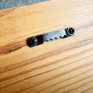 K&ouml;nnte beinhalten: Eine hellbraune Holzoberfl&auml;che mit einem schwarzen Metall-Zackenaufh&auml;nger, der mit zwei schwarzen Schrauben befestigt ist. Die Holzmaserung ist sichtbar und zeigt nat&uuml;rliche hell- und dunkelbraune Streifen, die horizontal verlaufen.