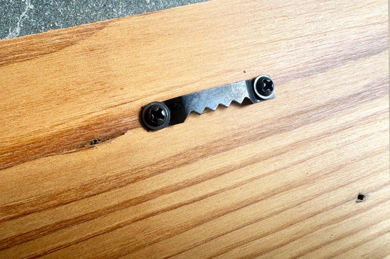 K&ouml;nnte beinhalten: Eine hellbraune Holzoberfl&auml;che mit einem schwarzen Metall-Zackenaufh&auml;nger, der mit zwei schwarzen Schrauben befestigt ist. Die Holzmaserung ist sichtbar und zeigt nat&uuml;rliche hell- und dunkelbraune Streifen, die horizontal verlaufen.