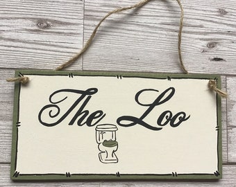 May include: A rectangular wooden sign with the words "The Loo" in black script, and a small toilet illustration. The sign has a green border and is suspended by a twine loop. The background is a light wood.