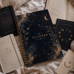May include: A spiral-bound planner with a dark blue cover featuring gold celestial motifs, stars, and foliage. The word PLANNER is printed in gold. It is surrounded by a cream-colored notebook, a black notebook with sun art, and a book showing constellations.