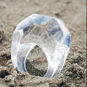 May include: A clear, geometric ring with a faceted design. The transparent ring is made of a clear material and sits on sand. The light reflects off the multiple flat surfaces of the ring, creating a sparkling effect.