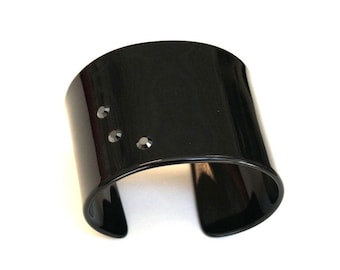 Op de afbeelding: Een brede, zwarte manchetarmband met een glanzende afwerking. Drie kleine, heldere kristallen zijn aan de voorkant ingebed. De armband heeft een open ontwerp. De sieraden zijn geplaatst tegen een witte achtergrond. De armband is ongeveer 5 cm breed.