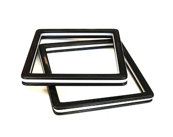 May include: Two square-shaped bangles featuring a layered design with a thick black outer border and a thin white center stripe. The bracelets have a smooth, glossy finish and are positioned overlapping each other to display their geometric form.