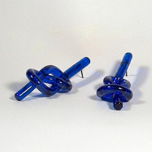 May include: A pair of cobalt blue glass stud earrings, each shaped into a knotted loop design. The earrings feature a smooth, translucent finish and a metal post backing, showcasing a vibrant, deep blue color throughout the knotted glass structure.