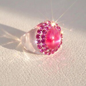 Op de afbeelding: Een heldere, dikke ring met een grote, roze, afgeronde middensteen. De middensteen is omgeven door een dubbele rij kleinere, roze edelstenen. De ring ligt op een wit oppervlak, met lichtreflectie op de facetten.