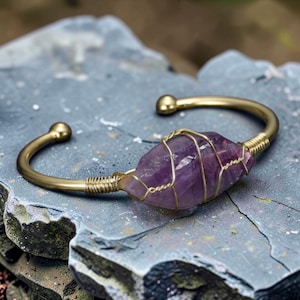 May include: A gold-toned metal cuff bracelet featuring a raw, purple amethyst crystal at its center. The crystal is secured to the open-ended band with thin, decorative gold-colored wire wrapping that extends along the sides of the stone.