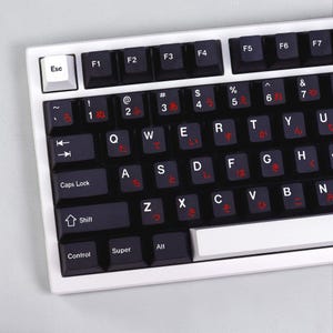 May include: A computer keyboard with a white frame and black keys. The keys feature white Latin alphabet letters and symbols, with additional red Japanese hiragana characters printed on each key. The Esc key is white, and the space bar is white.