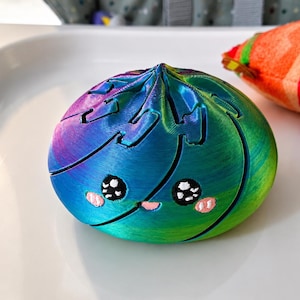 May include: A 3D printed dumpling-shaped fidget toy featuring a vibrant iridescent gradient of blue, purple, and green. The object has a smooth, rounded top with a pleated design and a printed face with black eyes and small pink cheeks.