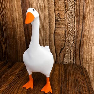 May include: A white goose figurine with a smooth matte finish, featuring a bright orange beak and matching orange webbed feet. The goose has a long, slender neck and small black eyes, standing in an upright, alert posture with a rounded body.
