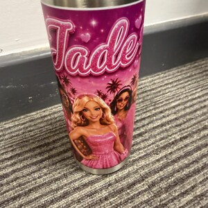 May include: A cylindrical tumbler with a black lid and a metallic rim. The exterior features a vibrant pink design with the name Jade in glittery script, small hearts, palm trees, and an illustration of three dolls in pink outfits.
