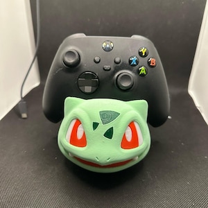 May include: A black wireless video game controller resting on a light green, 3D-printed stand shaped like the head of a Bulbasaur character. The controller features a central Xbox logo button, a directional pad, two analog sticks, and four colored action buttons.