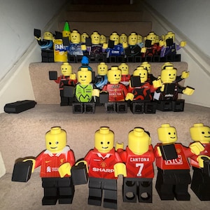 May include: A collection of yellow plastic minifigures arranged on stairs. The figures wear various sports jerseys, including red Manchester United shirts with Sharp and AIG logos, a red Liverpool jersey, and several Formula 1 racing team uniforms.