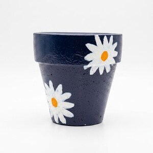 May include: A dark navy blue plant pot with a textured surface, featuring hand-painted white daisy flowers with bright yellow centers. The pot has a wide, flat rim and a tapered body, showcasing a simple floral design on the exterior.