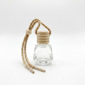 May include: A small, clear glass bottle with a geometric, faceted shape and a light-coloured wooden cap. The bottle features a beige, braided hanging cord with gold metallic threading and a single wooden bead for adjustment.