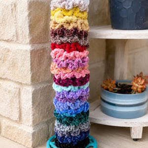 May include: A vertical stack of fifteen velvet hair scrunchies in a variety of colors including white, yellow, pink, red, purple, blue, green, gray, and black, arranged on a round, bright turquoise plastic stand with a circular cutout pattern.