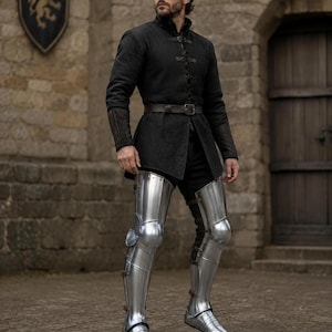 May include: A black long-sleeved tunic with a high collar and front lacing, worn with a dark brown leather belt and polished silver metal leg armor covering the thighs, knees, and shins, featuring articulated plates and pointed metal footwear.