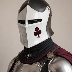 Op de afbeelding: Een metalen ridderhelm met een zwarte kap en een wit vizier voorzien van een donkerrood klaver-symbool. De helm heeft horizontale kijkspleten en een geperforeerd gedeelte, gedragen over een zwarte gewatteerde gambeson en zilveren schouderstukken.