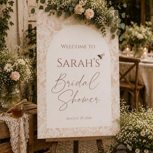 May include: A rectangular welcome sign for a bridal shower featuring an arched center panel with a beige lace pattern border. The text reads Welcome to Sarah's Bridal Shower August 29, 2026, with a small bee illustration. The sign is topped with roses and greenery.