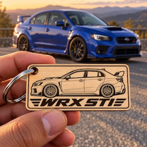 May include: A rectangular light-coloured wooden keyring featuring a laser-engraved side profile illustration of a saloon car with a rear spoiler and the text WRX STI printed in bold capital letters below the vehicle graphic, attached to a metal ring.