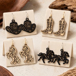 May include: Four pairs of laser-cut wooden earrings displayed on beige stands. The collection includes black cowboy hat shapes, light wood cowboy boot shapes, light wood rearing horse shapes, and black running horse shapes with intricate filigree.