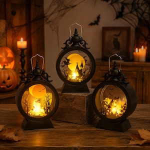 May include: Three circular black lanterns with metal handles, each featuring an illuminated interior scene with silhouettes of haunted houses, trees, bats, and a witch on a broomstick against a glowing yellow moon backdrop for Halloween decor.