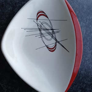 May include: A triangular ceramic dish with a white interior and a red outer rim. The center features a black and red graphic design consisting of intersecting lines, thin needles, and concentric oval shapes, creating a geometric pattern.