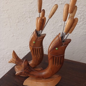 Op de afbeelding: Twee gesneden houten houders in de vorm van een vis, elk met een lichtbruine afwerking, kleine groene ogen en verticale kieuwgroeven. Elke vis bevat een set metalen aperitiefprikkers met bijpassende houten handvatten, rechtop in de open mond.