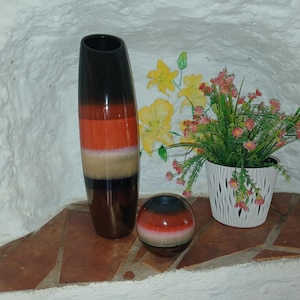 Könnte beinhalten: Eine hohe zylindrische Vase und eine kleine kugelförmige Vase, beide mit horizontalen Bändern aus dunkelbrauner, orangefarbener und cremefarbener Glasur. Daneben steht ein weißer Kunststofftopf mit kleinen rosa Blüten und grünen Stielen.