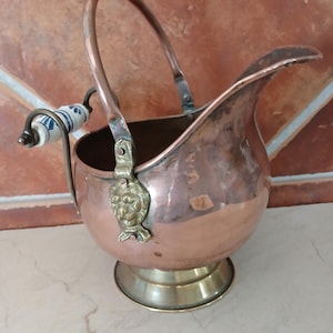 Pode incluir: Um jarro de cobre com base de latão e uma alça curva com um punho de cerâmica branca e azul. A alça é fixada ao corpo com suportes de latão em forma de cabeça de leão. O jarro tem um bico largo e um corpo arredondado de cobre polido.