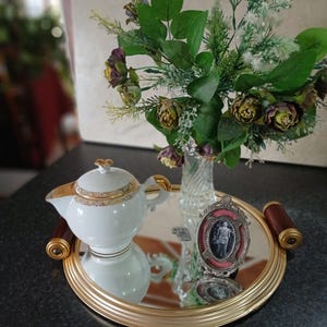 Peut inclure: Un plateau rond miroir à bordure dorée et poignées en bois foncé portant une théière en porcelaine blanche à liseré doré, un vase en verre transparent rempli de fleurs artificielles vertes et violettes, et un petit cadre photo ovale argenté.